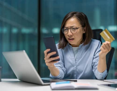 Mulher segurando celular e cartão de crédito, de frente para  o computador. 