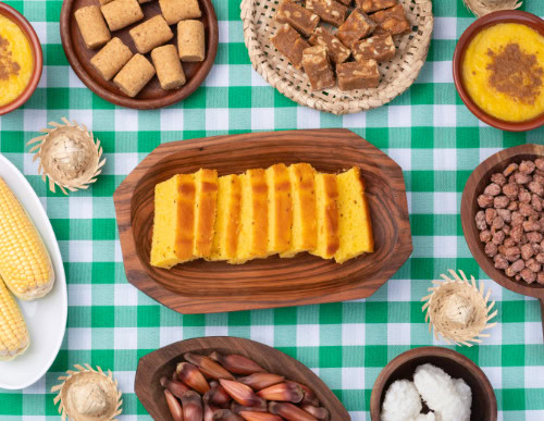 Mesa com pratos de Festa Junina: bolo fatiado, paçoca, pé de moça, pinhão, etc.
