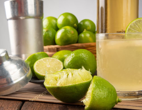 Limões cortados e inteiros, coqueteleira e copo com caipirinha.