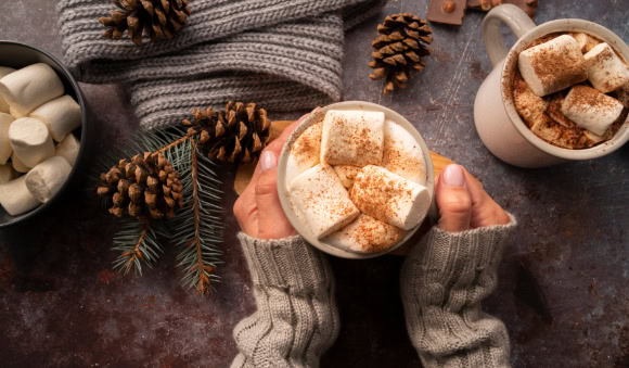 Mãos segurando copo com chocolate quente, coberto por chantilly e marshmallow.