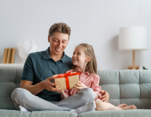 Pai e filha sentados no sofá. Pai segura presente. 