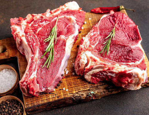 Dois pedaços de carne sobre tábua de madeira com ramo de alecrim por cima.