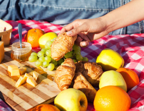 Mão segurando croissant em tábua de frios e frutas em piquenique.